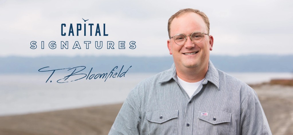 A smiling man wearing glasses and a striped shirt stands in front of a scenic coastal background, with 'Capital Signatures' and 'T. Bloomfield' text overlay.