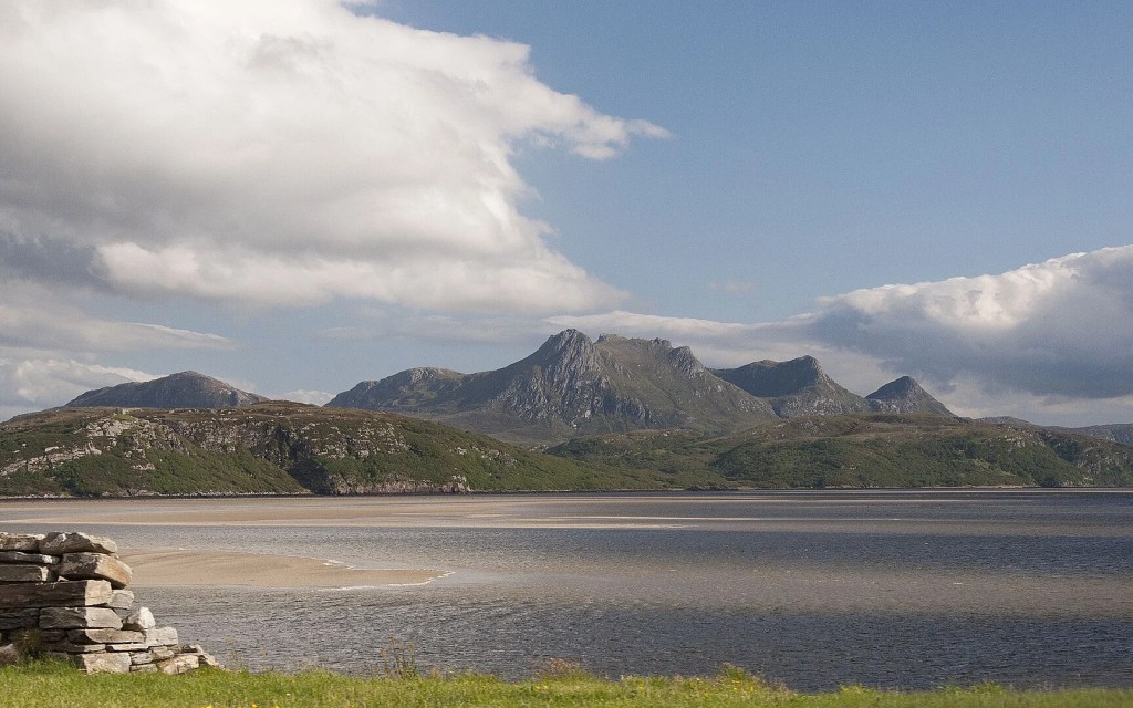 A scenic view of the Kyle of Tongue sea loch in northwest Highland, Scotland, featuring the distinctive peaks of Ben Loyal mountain in the background. The shallow waters of the sea loch stretch across the foreground, with tidal flats visible during low tide. The landscape shows typical Highland terrain with rocky shores, green vegetation, and the characteristic moody Scottish sky overhead. The natural beauty of this National Scenic Area is captured in the interplay between water, mountains, and sky