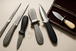 A high-resolution editorial photograph of several types of oyster knives that should be avoided, including long flexible blades, dull rounded-tip knives, smooth plastic-handled knives, overly sharp razor-edged blades, and decorative boxed gift-set knives. The tools are arranged on a clean, neutral background to emphasize the differences and highlight safety concerns.