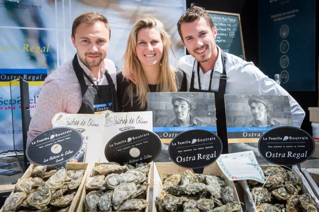 Three people stand behind a stall displaying baskets of Ostra Regal oysters and La Famille Boutrais signage at a seafood market