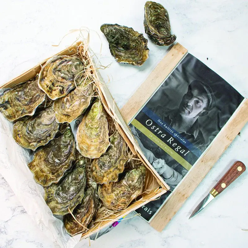 A wooden crate of graded Ostra Regal oysters beside branded packaging and a small oyster knife