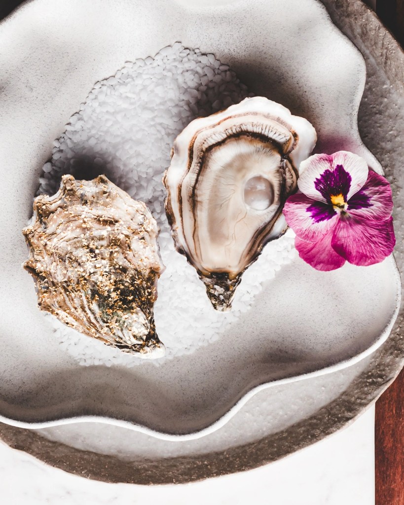  Two Ostra Regal oysters, one shucked and one whole, presented in a bowl of sea salt with a decorative edible flower.