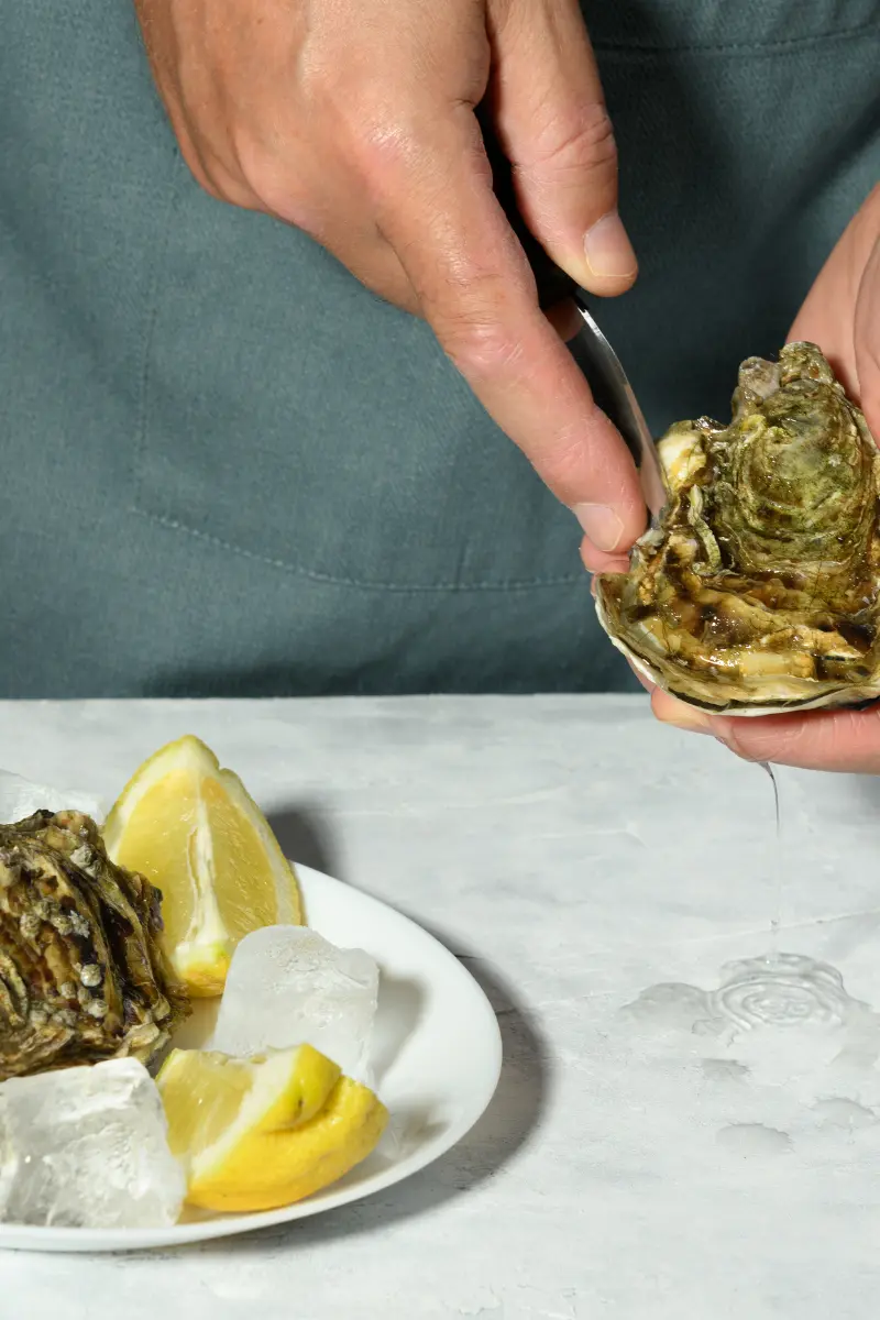 Wide view of oyster shucking with the oyster held low over the counter and a plated oyster on ice nearby